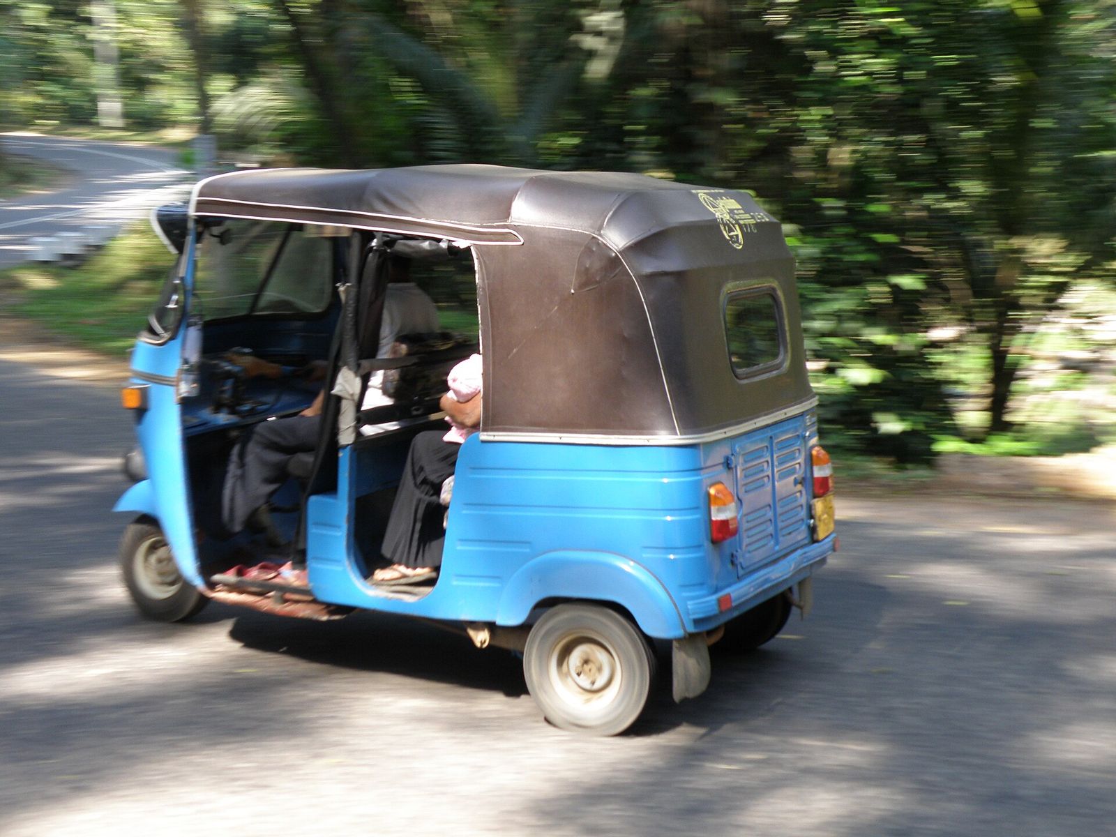 A blue tuk tuk speeding along a tree-lined road